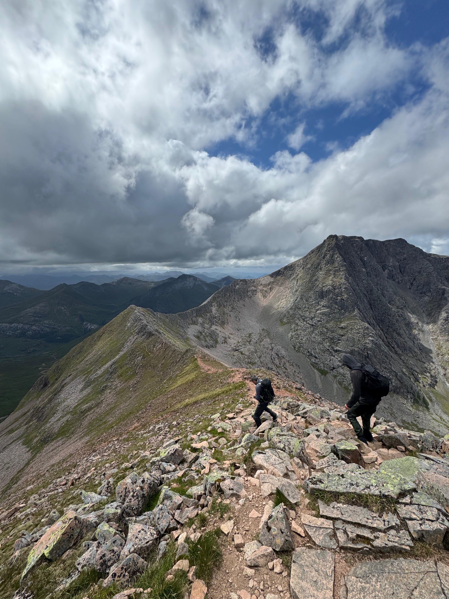 Two hikers descending a rocky mountain ridge beneath dramatic clouds