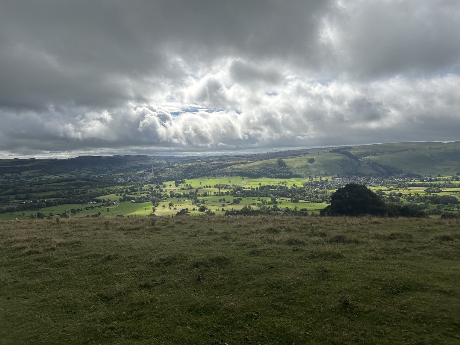 Sunlight breaking through the clouds onto green fields and wooded hills in the Peak District