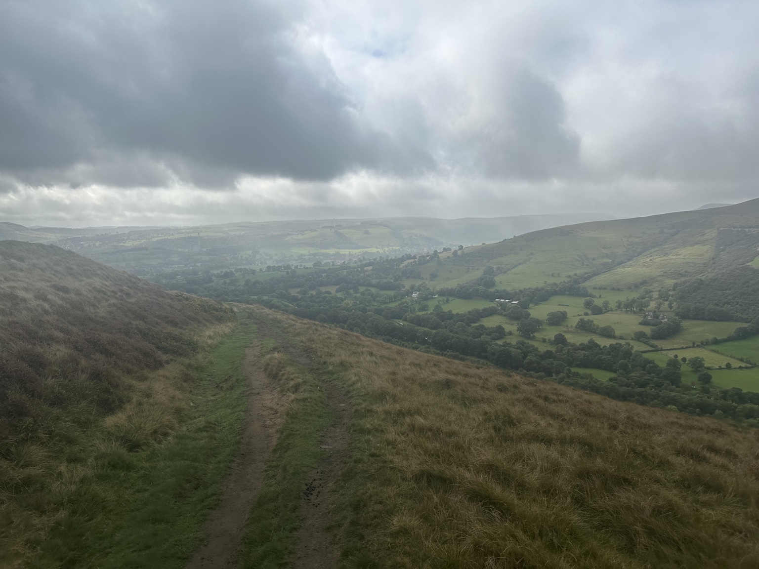 A path across the grassy hillside above the valley beneath low grey cloud